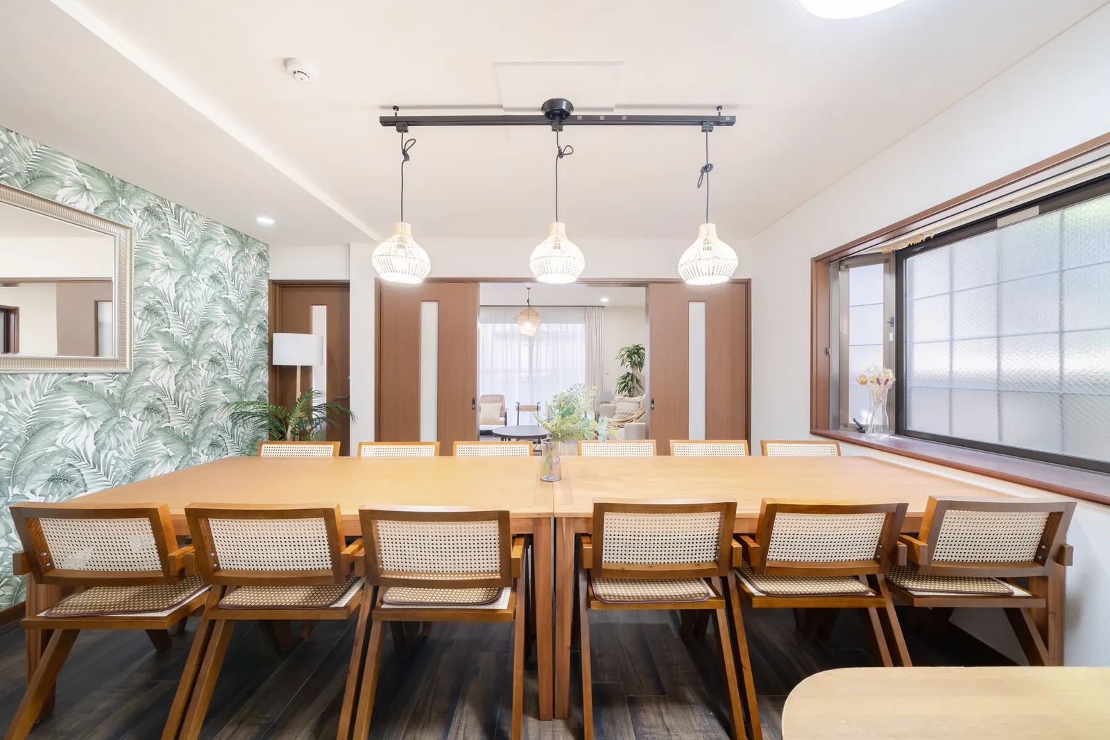 Long dining table with twelve cane chairs under three rattan pendant lamps at Hotel MADO Shin-Okubo, with a palm-leaf feature wall and the living room visible beyond