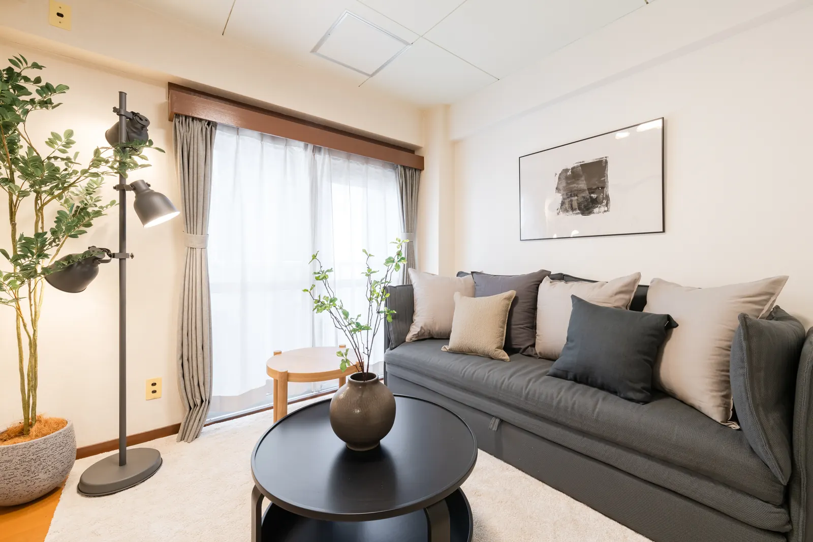 Living room of the apartment with a grey three-seater sofa that converts to a double bed, cushions, a black round table, a floor lamp and a balcony window